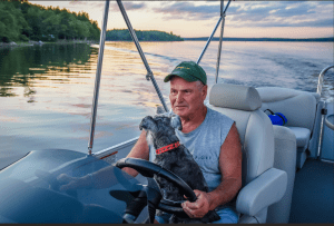 A senior man drives a pontoon with a dog in his lap.