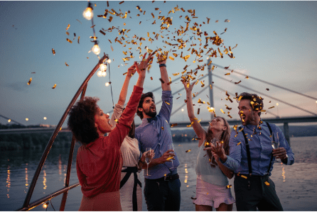 A group of men and women throwing confetti up into the air with water and a bridge in the background. 