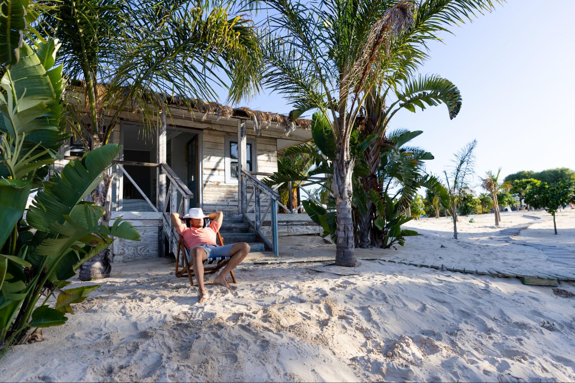 A young Caucasian man sits on a deck chair outside a rustic cottage, his hat covering his face. The setting is serene, surrounded by palm trees and white sand at the beach.