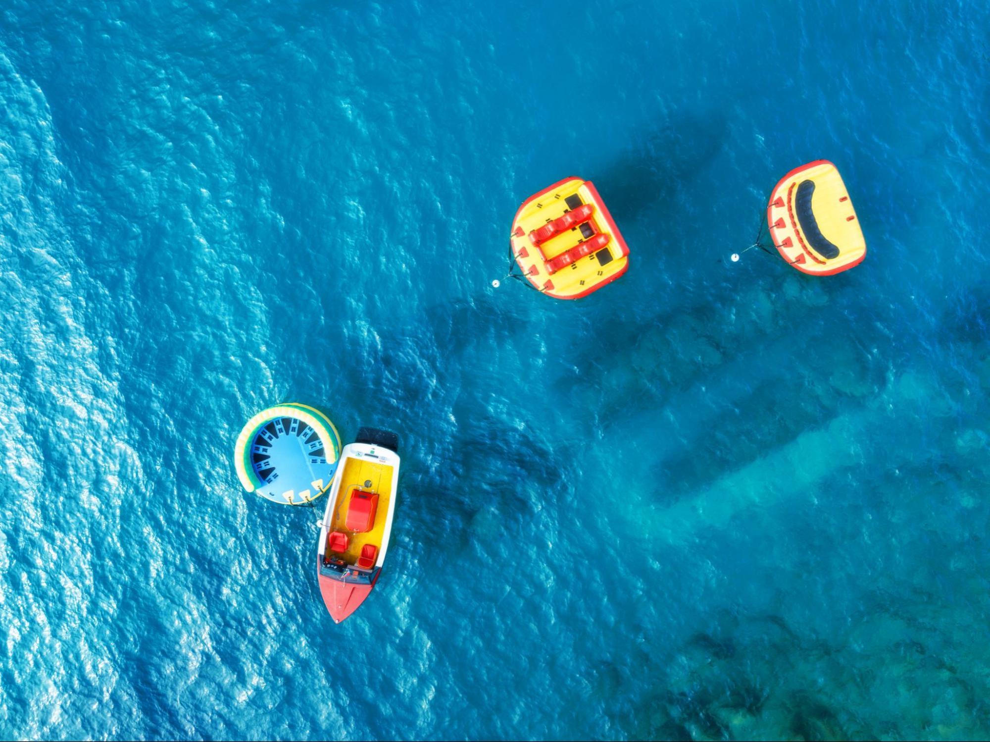 An aerial view of a boat towing a fly fish inflatable ring with two other yellow and orange inflatables awaiting water adventure seekers on a clear blue sea.