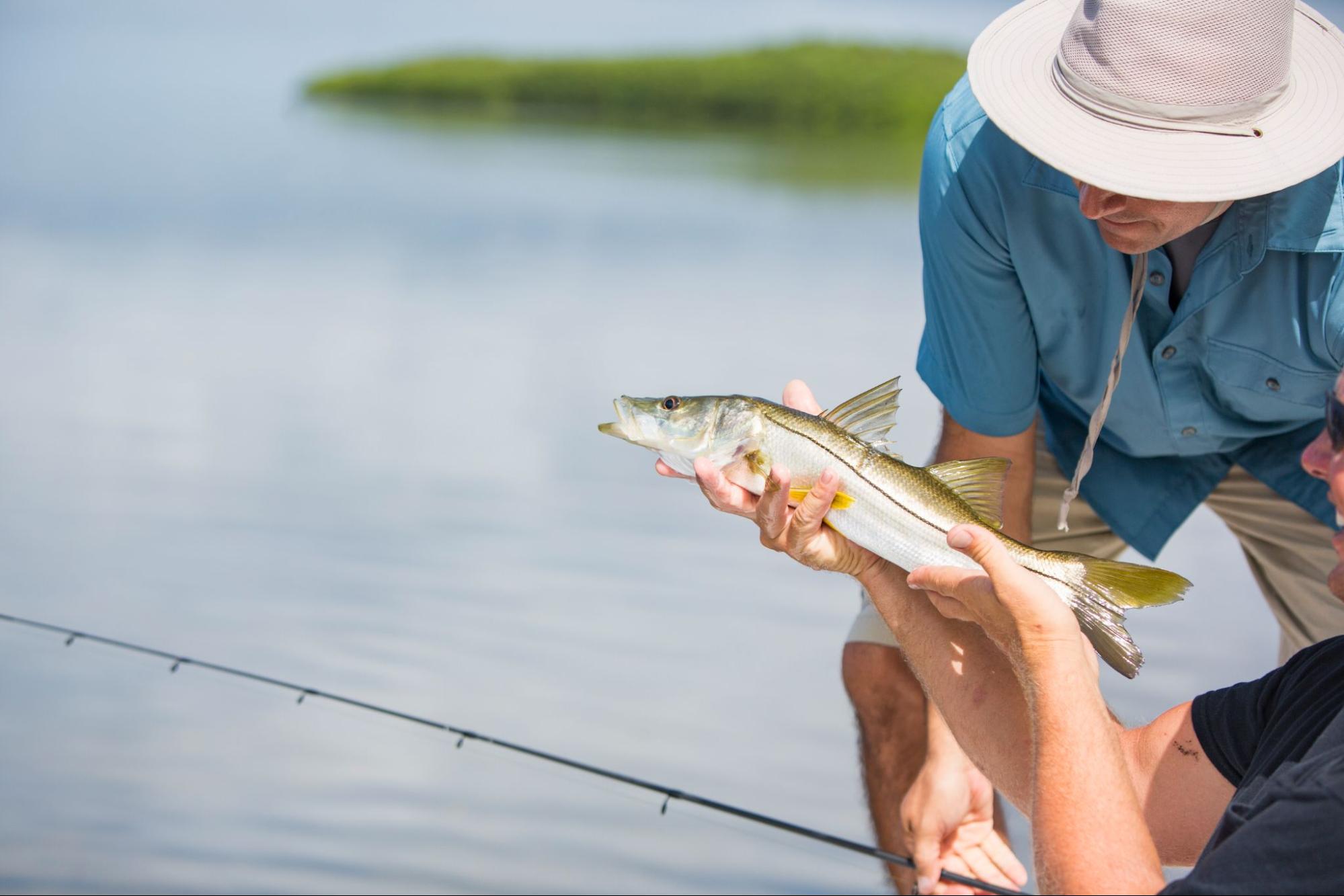 Two men fishing with one holding a fish next to open water. 