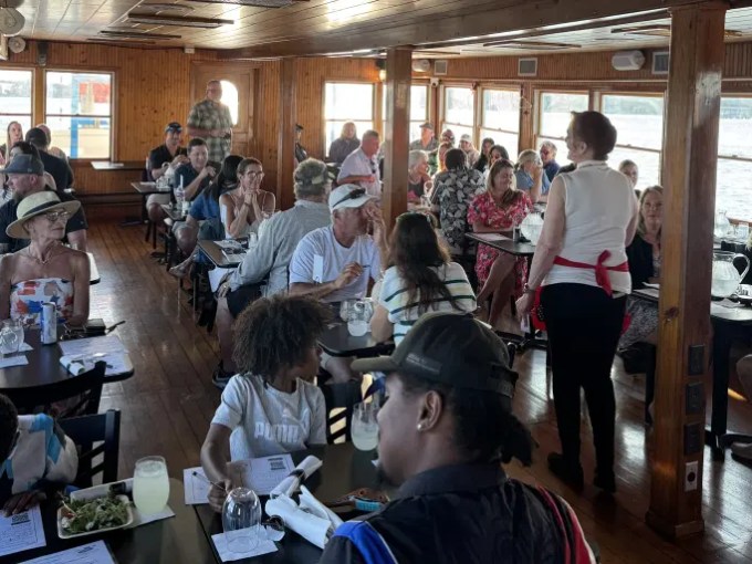 People dining in a wood-paneled boat interior with large windows.